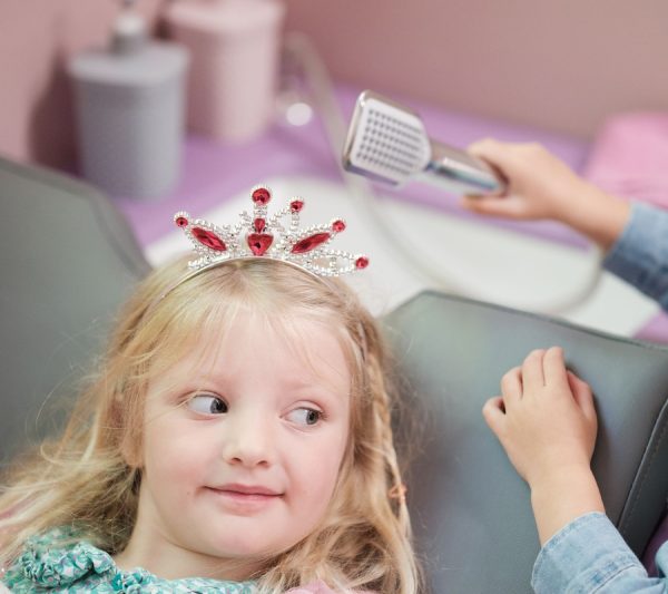 A little girl wearing a princess tiara, laying in the salon chair at Jack in the Box, while another child brushes her hair as part of their role play.